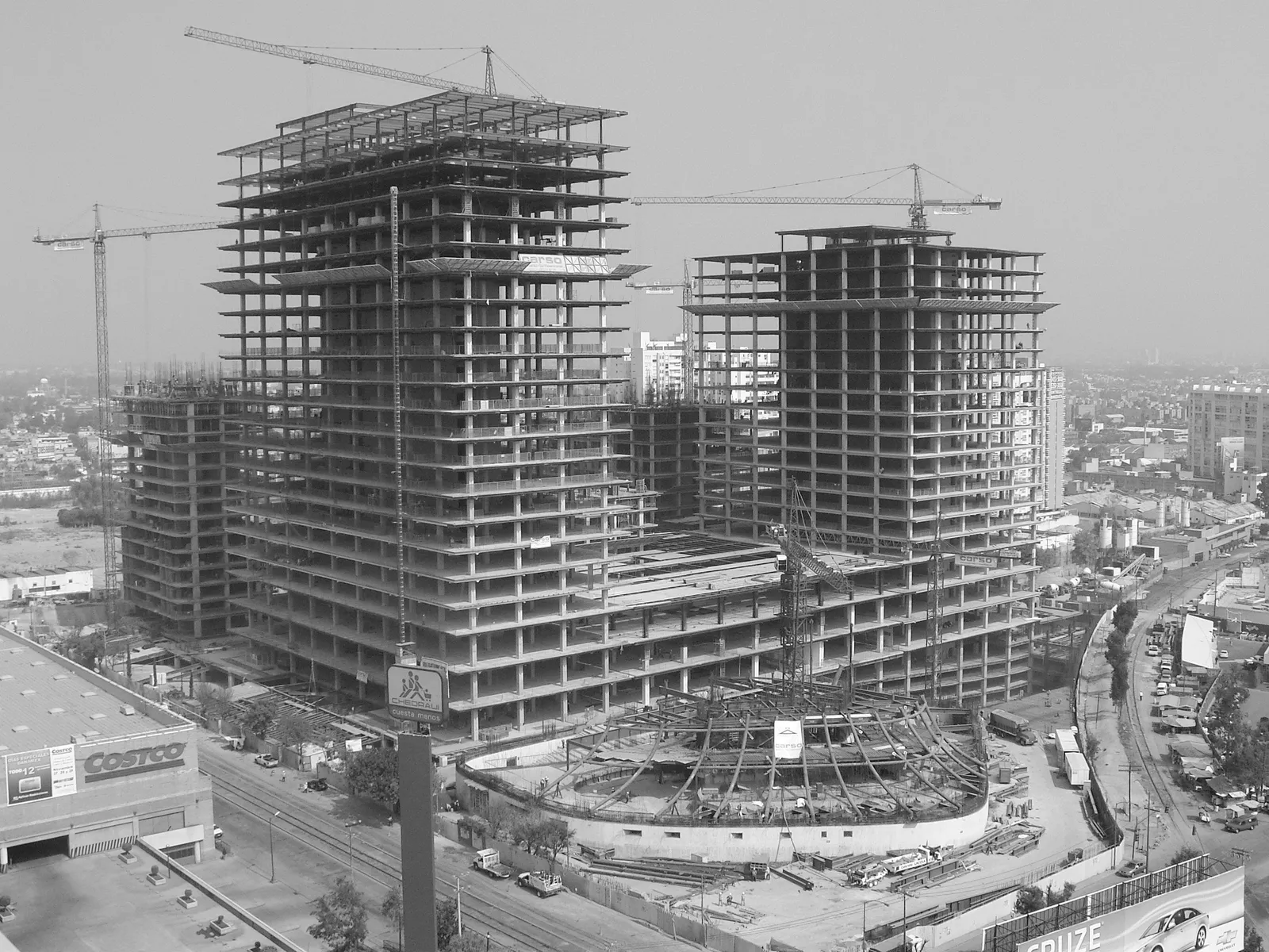 Plaza Carso under construction: two towers with exposed steel framing and cranes, the city in the background.
